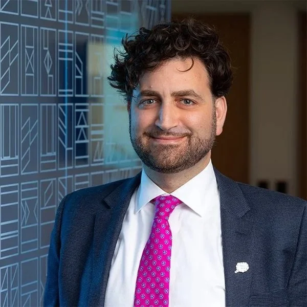 Smiling man in a suit with curly hair, a pink patterned tie, and a lapel pin, stands against a geometric-patterned wall, conveying confidence.