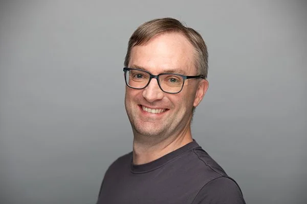 Smiling man wearing glasses and a dark shirt against a gray background. The tone is friendly and approachable.