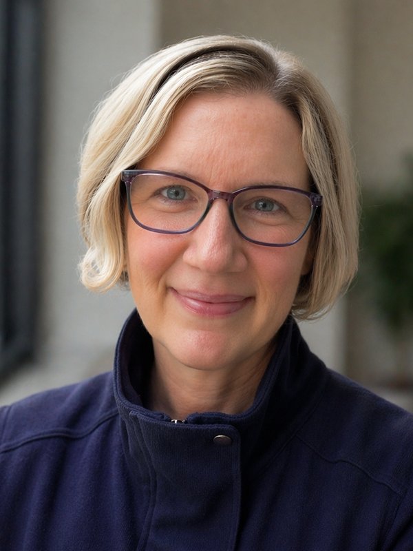 A person with short, light blonde hair, wearing glasses and a navy jacket, smiles warmly at the camera. The background is softly blurred, suggesting an indoor setting.
