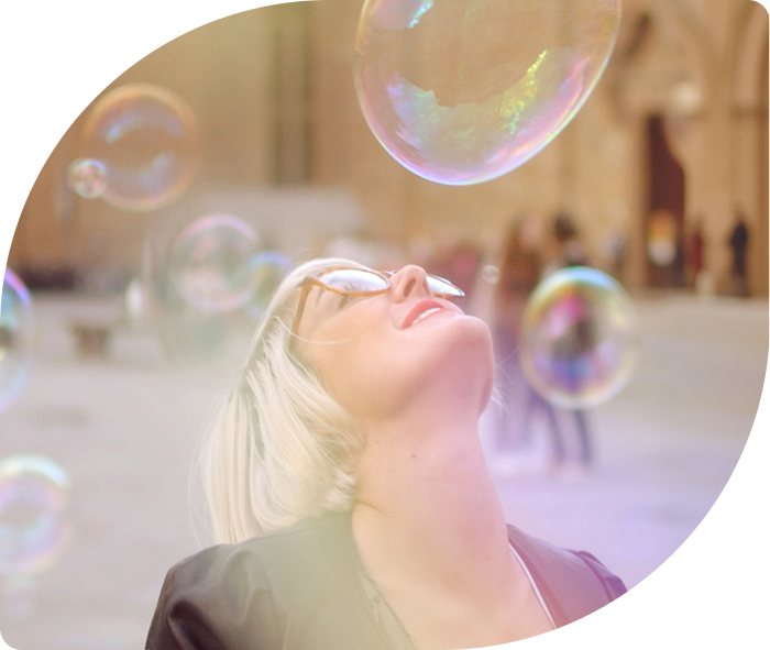 A person wearing sunglasses looking up at large soap bubbles floating in an outdoor plaza