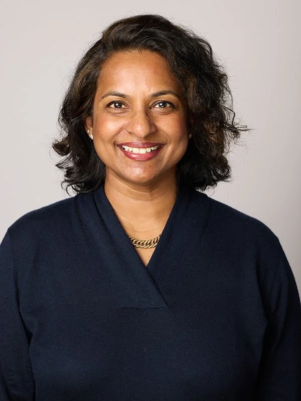 A woman smiles warmly, wearing a dark blue top and a gold necklace, against a light, neutral background. The tone is friendly and approachable.