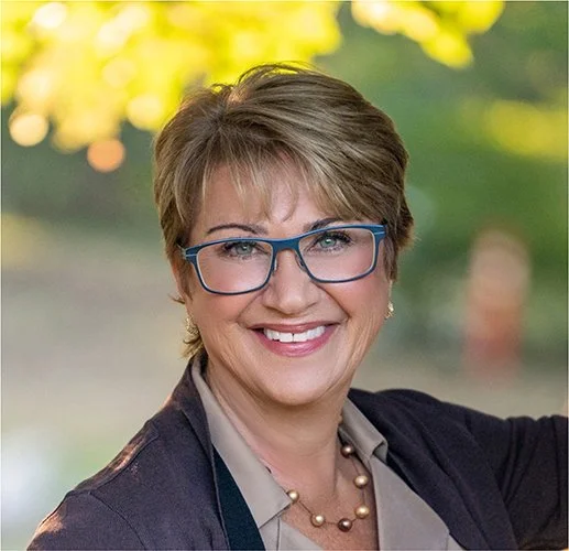 A woman with short hair and blue glasses smiles warmly. She wears a blazer, a blouse, and a beaded necklace. Blurred green and yellow background.