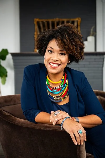 Smiling woman with curly hair wearing a navy blazer, colorful beaded necklaces, and rings, sitting on a brown sofa. The atmosphere is warm and inviting.