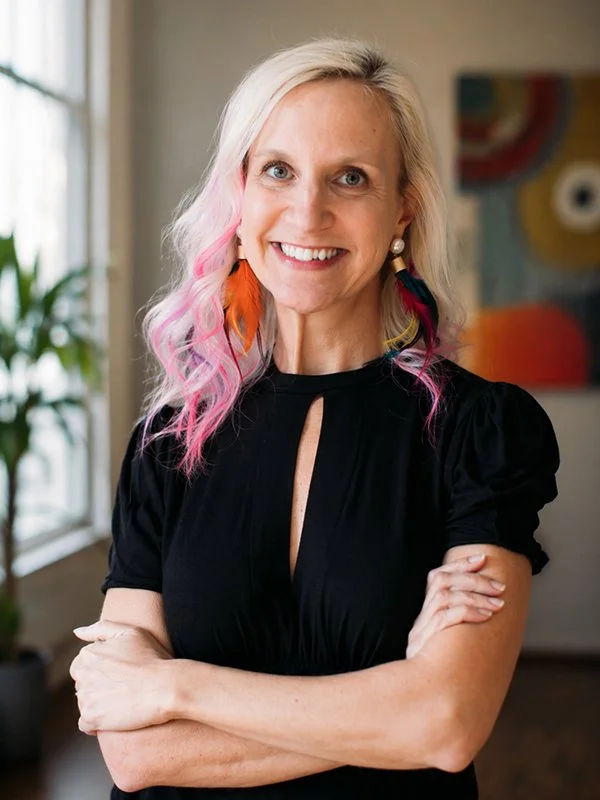 Smiling woman with pink and purple hair highlights wears colorful feather earrings and a black dress, standing in a bright room with a plant.