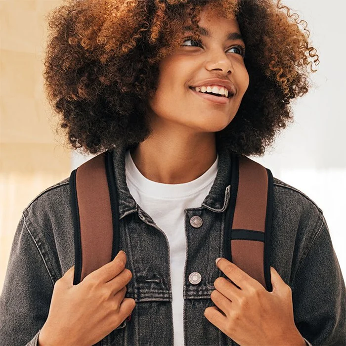 A smiling young person wearing a denim jacket and a brown backpack