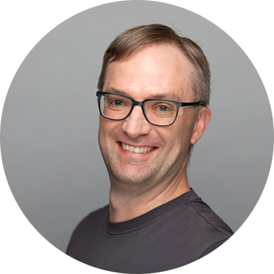 Smiling man wearing glasses and a dark shirt against a gray background. The tone is friendly and approachable.