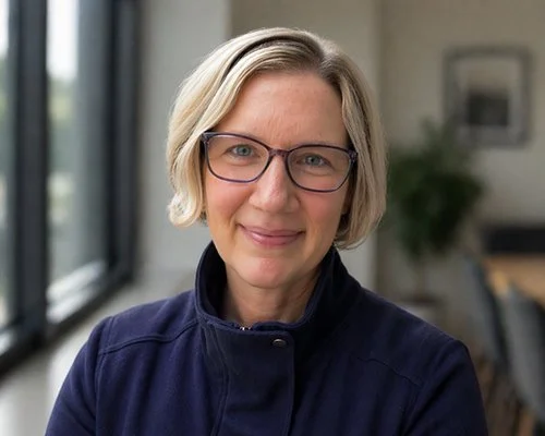 A person with short, light blonde hair, wearing glasses and a navy jacket, smiles warmly at the camera. The background is softly blurred, suggesting an indoor setting.