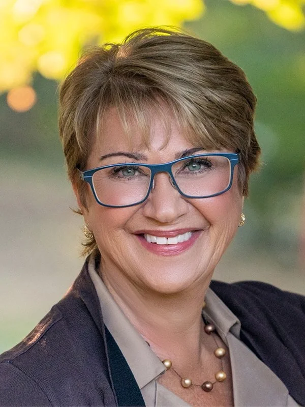 A woman with short hair and blue glasses smiles warmly. She wears a blazer, a blouse, and a beaded necklace. Blurred green and yellow background.