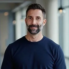 Man with a beard and short dark hair smiling warmly, wearing a navy sweater, standing in a bright hallway with soft lighting. Calm and friendly mood.
