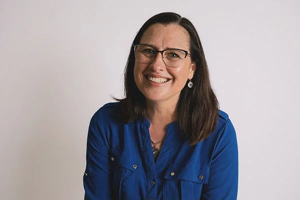 Smiling woman with glasses, wearing a blue shirt and earrings, against a plain white background. The tone is cheerful and welcoming.