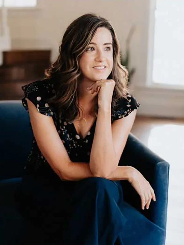 A woman with wavy hair sits on a blue sofa, resting her chin on her hand, smiling thoughtfully. She's wearing a dark, patterned dress. Bright, cozy room.