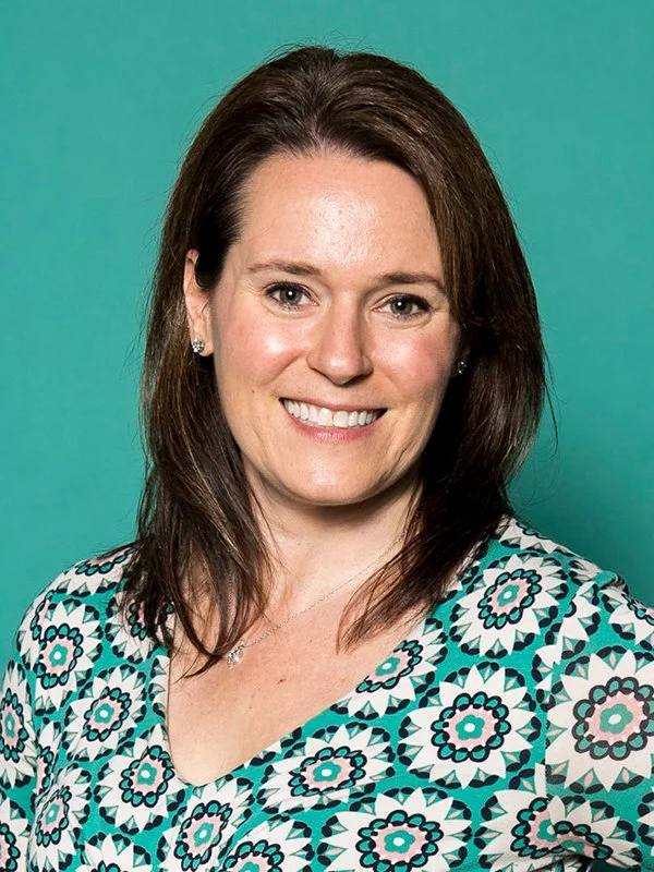 Smiling woman with brown hair, wearing a patterned teal and black blouse, against a solid teal background. The mood is friendly and approachable.