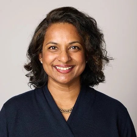 A woman smiles warmly, wearing a dark blue top and a gold necklace, against a light, neutral background. The tone is friendly and approachable.