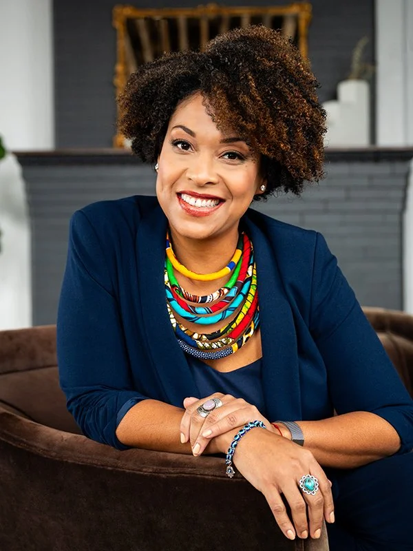 Smiling woman with curly hair wearing a navy blazer, colorful beaded necklaces, and rings, sitting on a brown sofa. The atmosphere is warm and inviting.