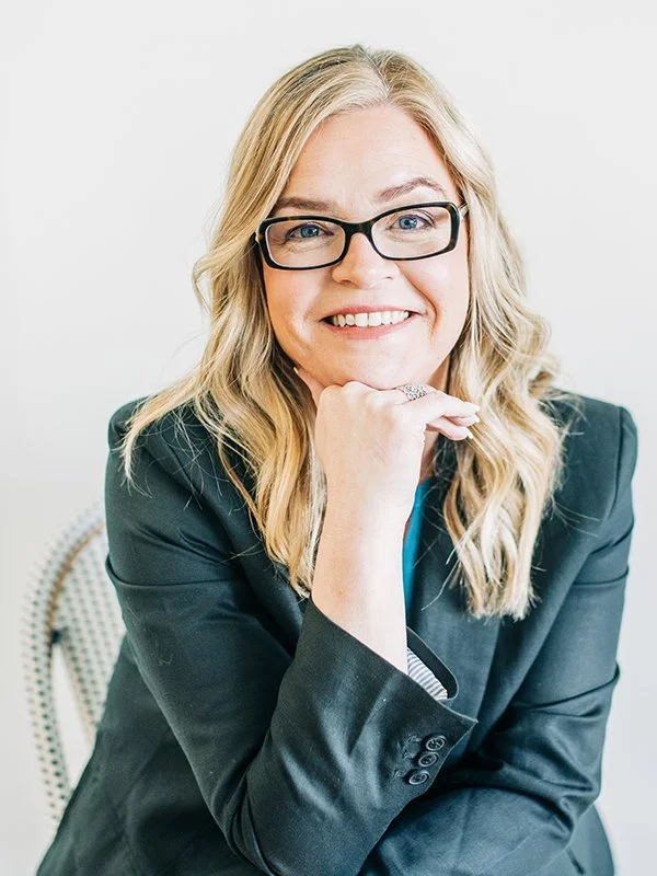 A smiling woman with blonde hair and glasses sitting in a chair, wearing a dark blazer. She rests her chin on her hand, conveying a friendly demeanor