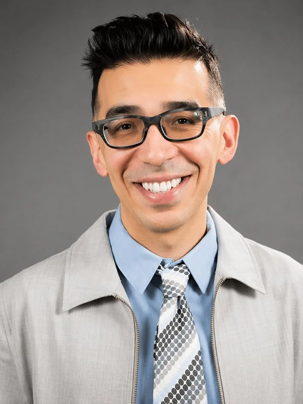 Smiling man with glasses and short dark hair, wearing a light gray blazer, blue shirt, and patterned tie. Gray background. Professional and cheerful tone.