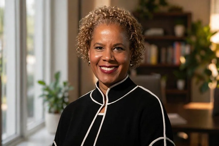 Smiling woman with short curly hair wearing a black top with white trim. Background includes bookshelves with blurred books, creating a professional, warm tone.