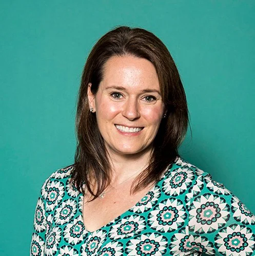 Smiling woman with brown hair, wearing a patterned teal and black blouse, against a solid teal background. The mood is friendly and approachable.