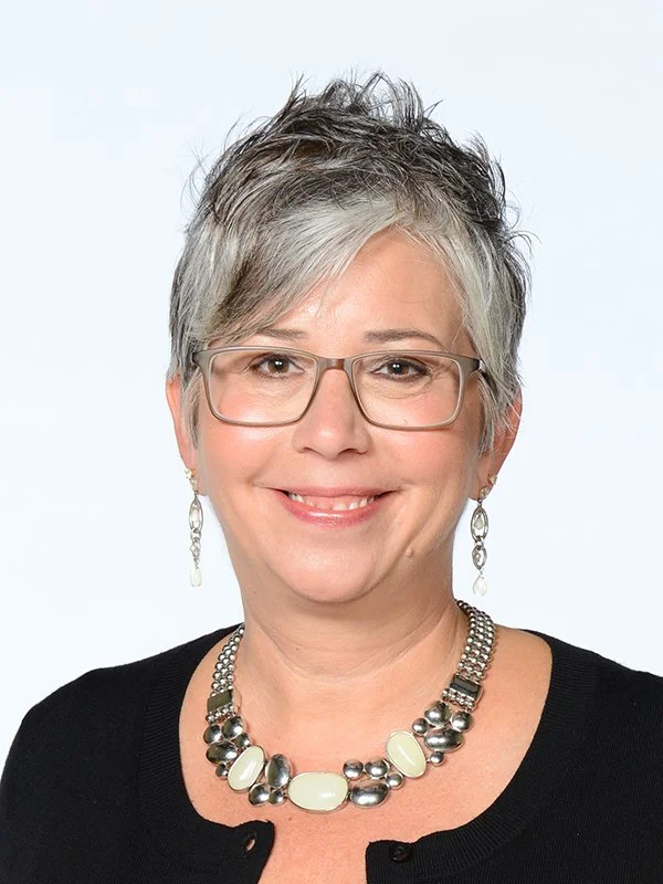 A woman with short gray hair smiles warmly at the camera. She wears glasses, a black top, dangling earrings, and a chunky beaded necklace, exuding confidence.