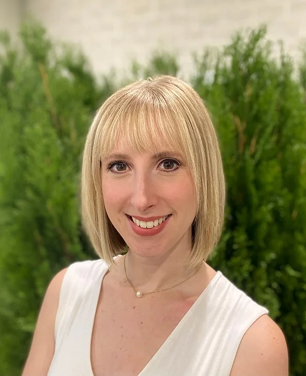 A woman with short blonde hair and bangs smiles warmly against a backdrop of green foliage. She wears a white top and a thin necklace with a pearl pendant.