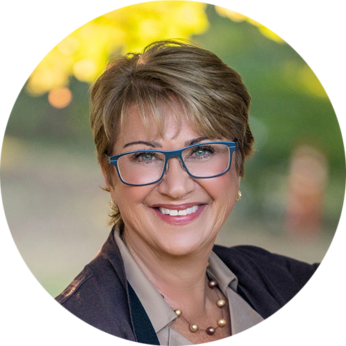 A woman with short hair and blue glasses smiles warmly. She wears a blazer, a blouse, and a beaded necklace. Blurred green and yellow background.