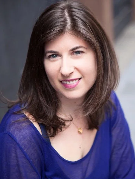 A woman with shoulder-length brown hair and a friendly smile wears a blue top. The background is softly blurred, emphasizing her warm, approachable expression.
