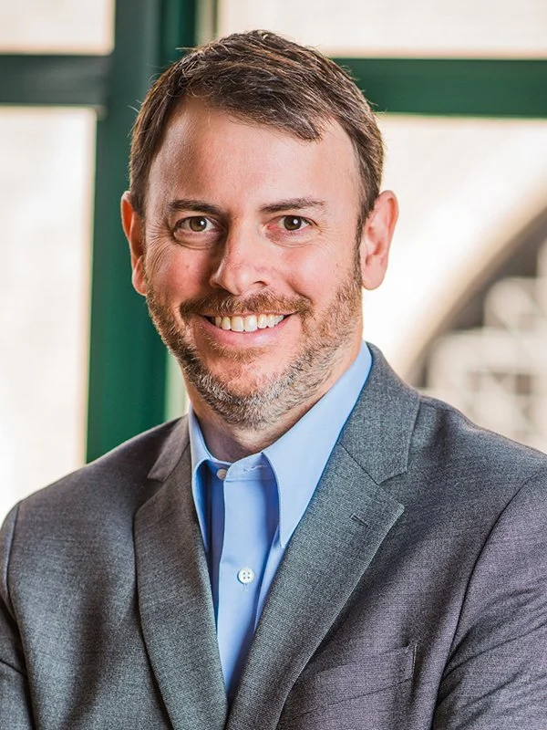 A smiling man with short hair and a beard is wearing a gray suit and blue shirt. He stands indoors, with a blurred window behind him.