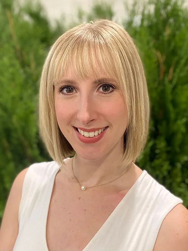 A woman with short blonde hair and bangs smiles warmly against a backdrop of green foliage. She wears a white top and a thin necklace with a pearl pendant.