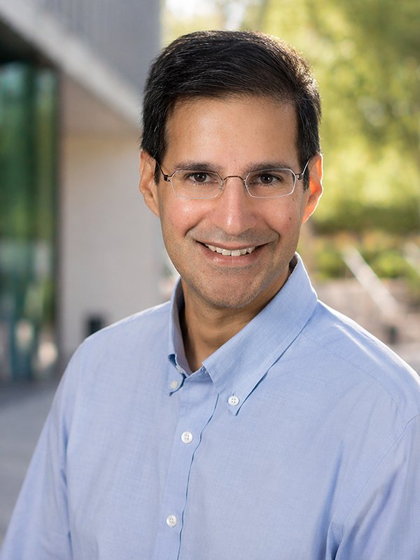 Smiling man wearing glasses and a light blue shirt stands outdoors with greenery and building in the background, conveying a friendly tone.