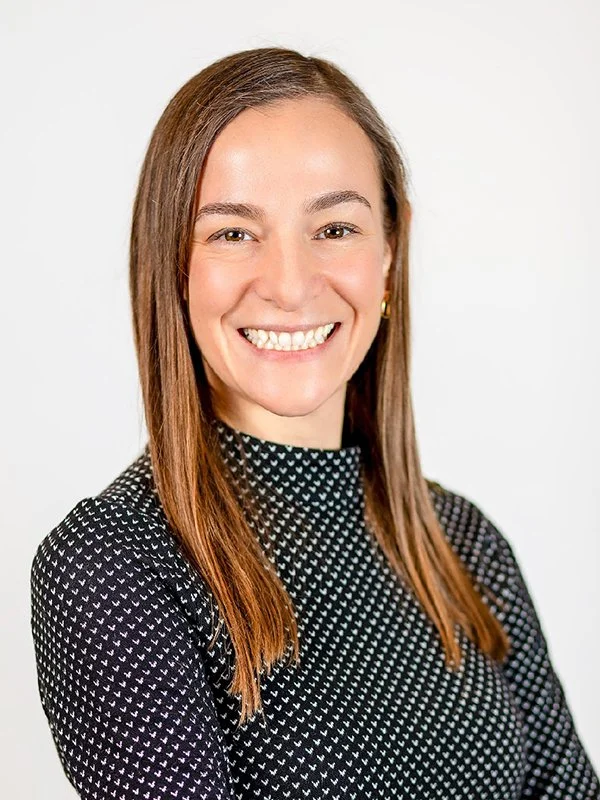Smiling woman with long brown hair, wearing a black patterned sweater. She stands against a plain white background, exuding a friendly and confident demeanor.