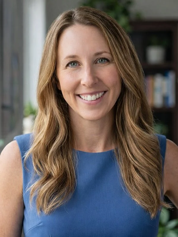 A woman with long, wavy blonde hair smiles warmly, wearing a blue sleeveless top. She stands indoors with bookshelves in the softly focused background.