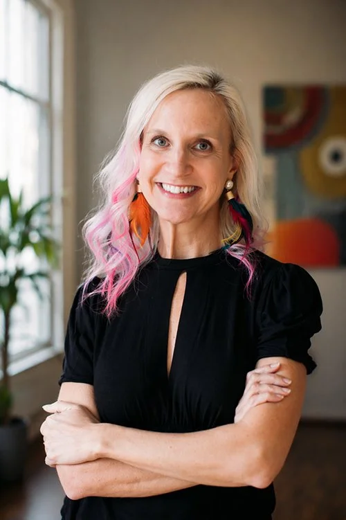 Smiling woman with pink and purple hair highlights wears colorful feather earrings and a black dress, standing in a bright room with a plant.