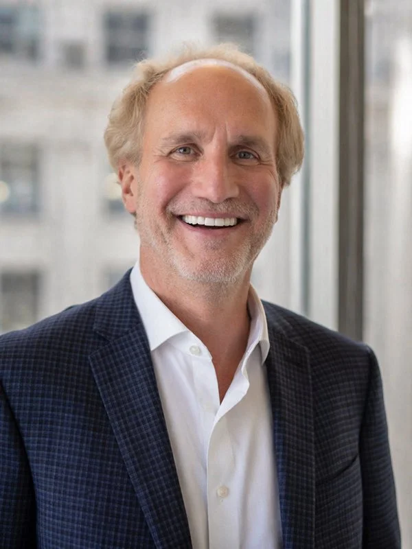 A smiling man with short, light hair is wearing a blue checkered suit and white shirt against a blurred cityscape through a window background.