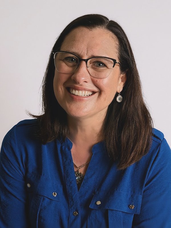 Smiling woman with glasses, wearing a blue shirt and earrings, against a plain white background. The tone is cheerful and welcoming
