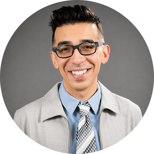Smiling man with glasses and short dark hair, wearing a light gray blazer, blue shirt, and patterned tie. Gray background. Professional and cheerful tone.