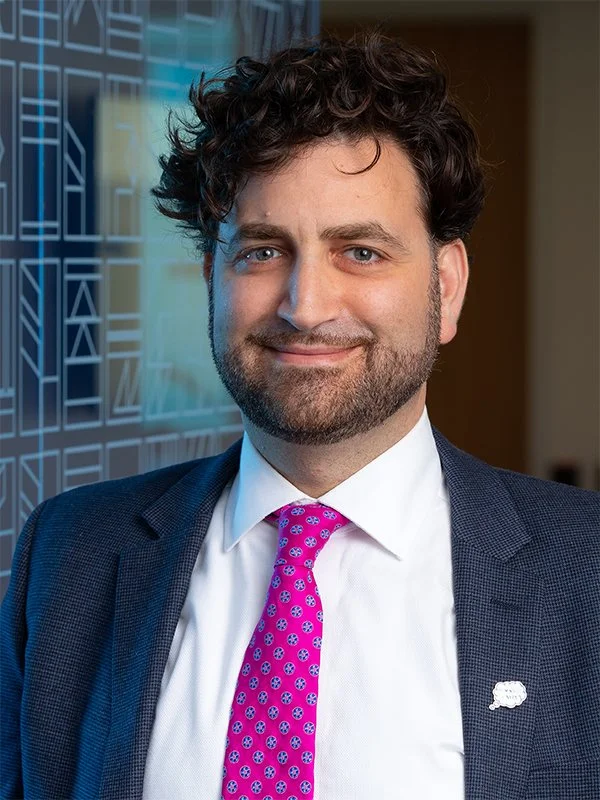 Smiling man in a suit with curly hair, a pink patterned tie, and a lapel pin, stands against a geometric-patterned wall, conveying confidence.
