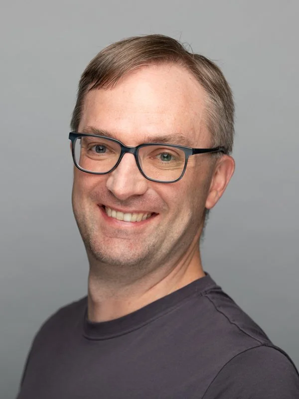 Smiling man wearing glasses and a dark shirt against a gray background. The tone is friendly and approachable.