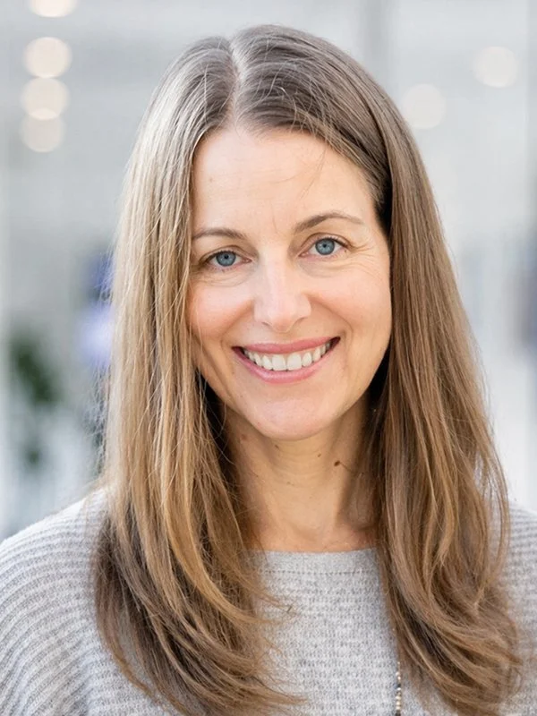 Smiling woman with long brown hair wearing a gray sweater. Soft, light-filled background with blurred lights, conveying warmth and friendliness.