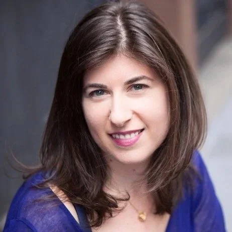 A woman with shoulder-length brown hair and a friendly smile wears a blue top. The background is softly blurred, emphasizing her warm, approachable expression.