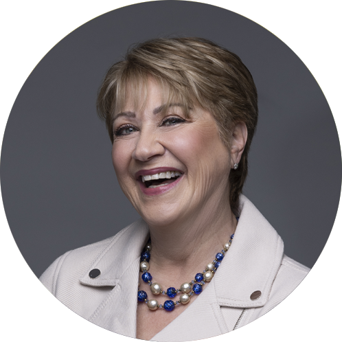 A woman with short hair and blue glasses smiles warmly. She wears a blazer, a blouse, and a beaded necklace. Blurred green and yellow background.