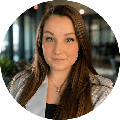 A young woman with long brown hair and blue eyes looks into the camera. She wears a light-colored jacket and a delicate necklace, with a calm expression. Blurred background.
