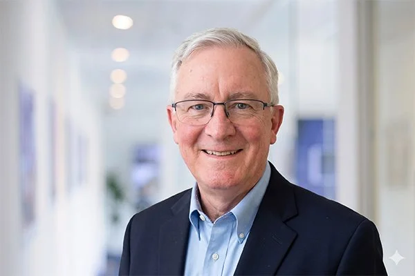 Smiling older man with glasses and short white hair, wearing a blue shirt and dark blazer. The background is a softly blurred office setting.