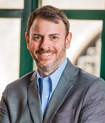 A smiling man with short hair and a beard is wearing a gray suit and blue shirt. He stands indoors, with a blurred window behind him.