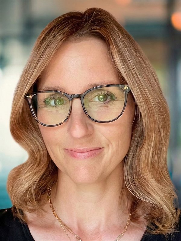 A woman with wavy brown hair and glasses smiles softly. She wears a black top and gold necklace. The background is softly blurred, creating a warm, inviting tone.