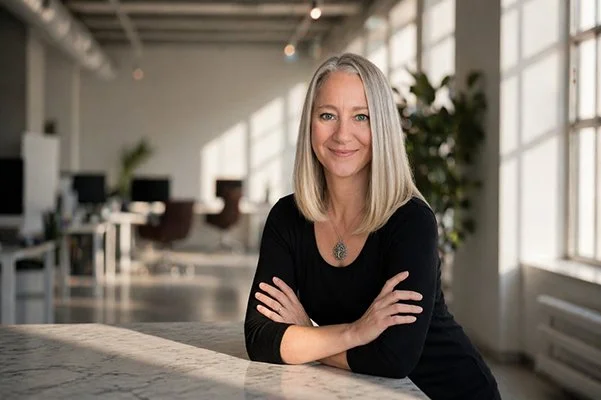 A woman with shoulder-length blonde hair smiles confidently, arms crossed, wearing a black top and pendant. Bright, modern room in the background.