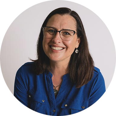 Smiling woman with glasses, wearing a blue shirt and earrings, against a plain white background. The tone is cheerful and welcoming.
