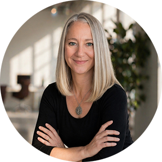 A woman with shoulder-length blonde hair smiles confidently, arms crossed, wearing a black top and pendant. Bright, modern room in the background.