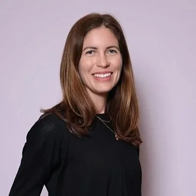A smiling woman with long brown hair wearing a black top and necklace poses in front of a plain light purple background, conveying a friendly and approachable tone.