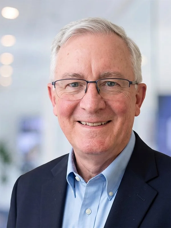 Smiling older man with glasses and short white hair, wearing a blue shirt and dark blazer. The background is a softly blurred office setting.