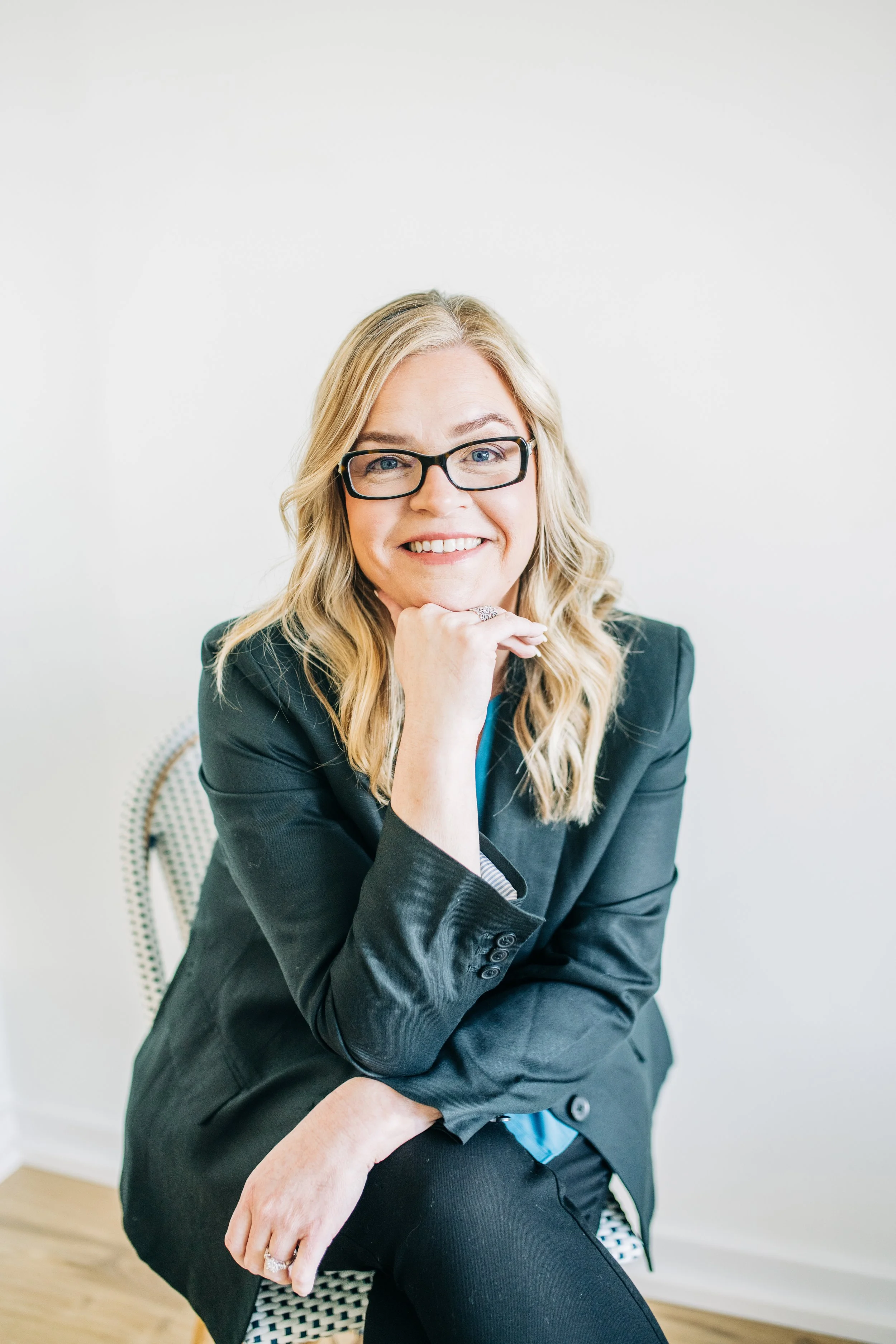 A smiling woman with blonde hair and glasses sitting in a chair, wearing a dark blazer. She rests her chin on her hand, conveying a friendly demeanor.